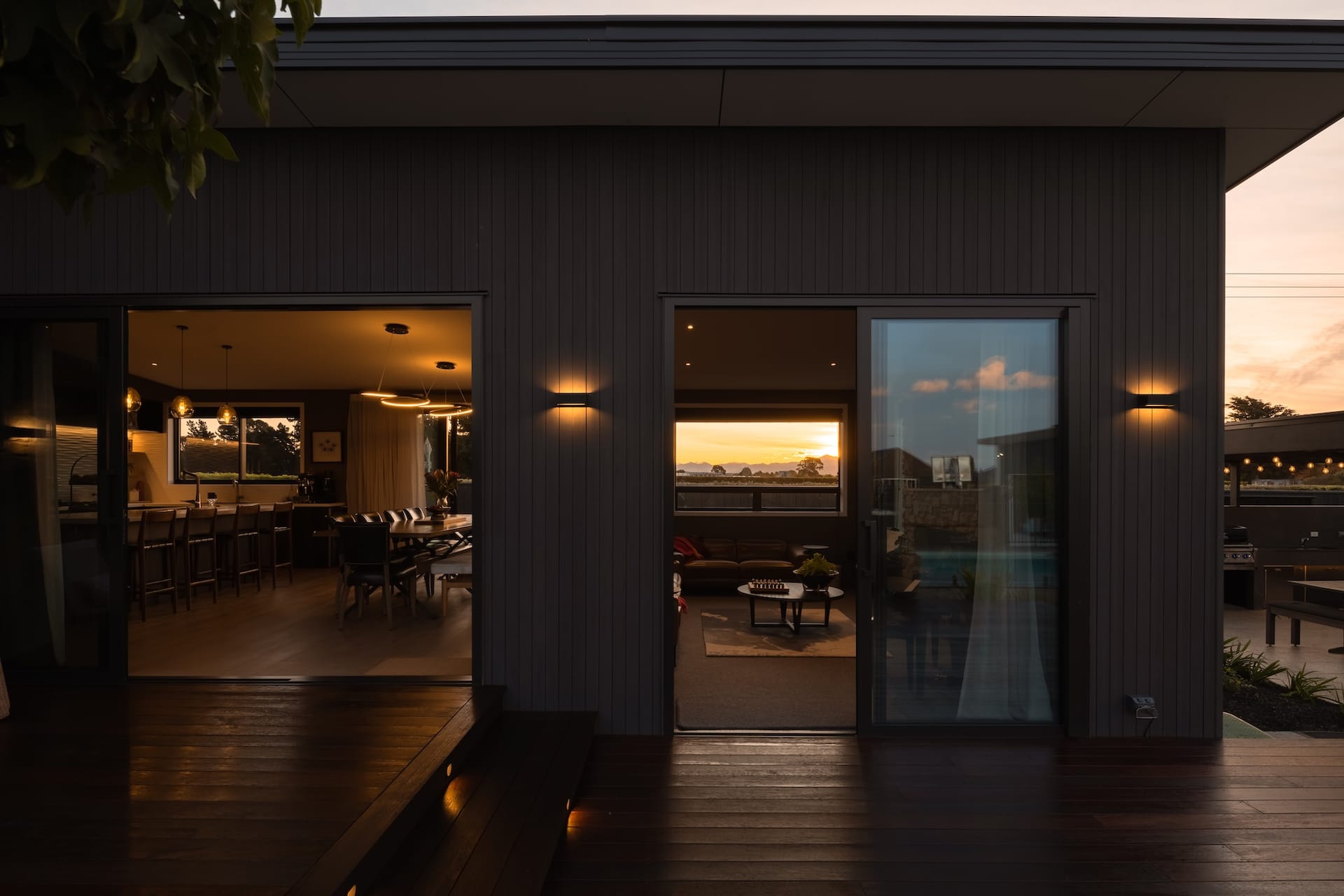A Discovery Homes new build at Spitfire Drive at dusk, dark cladding with warm interior light spilling through open stacker doors and a sunset over the valley beyond
